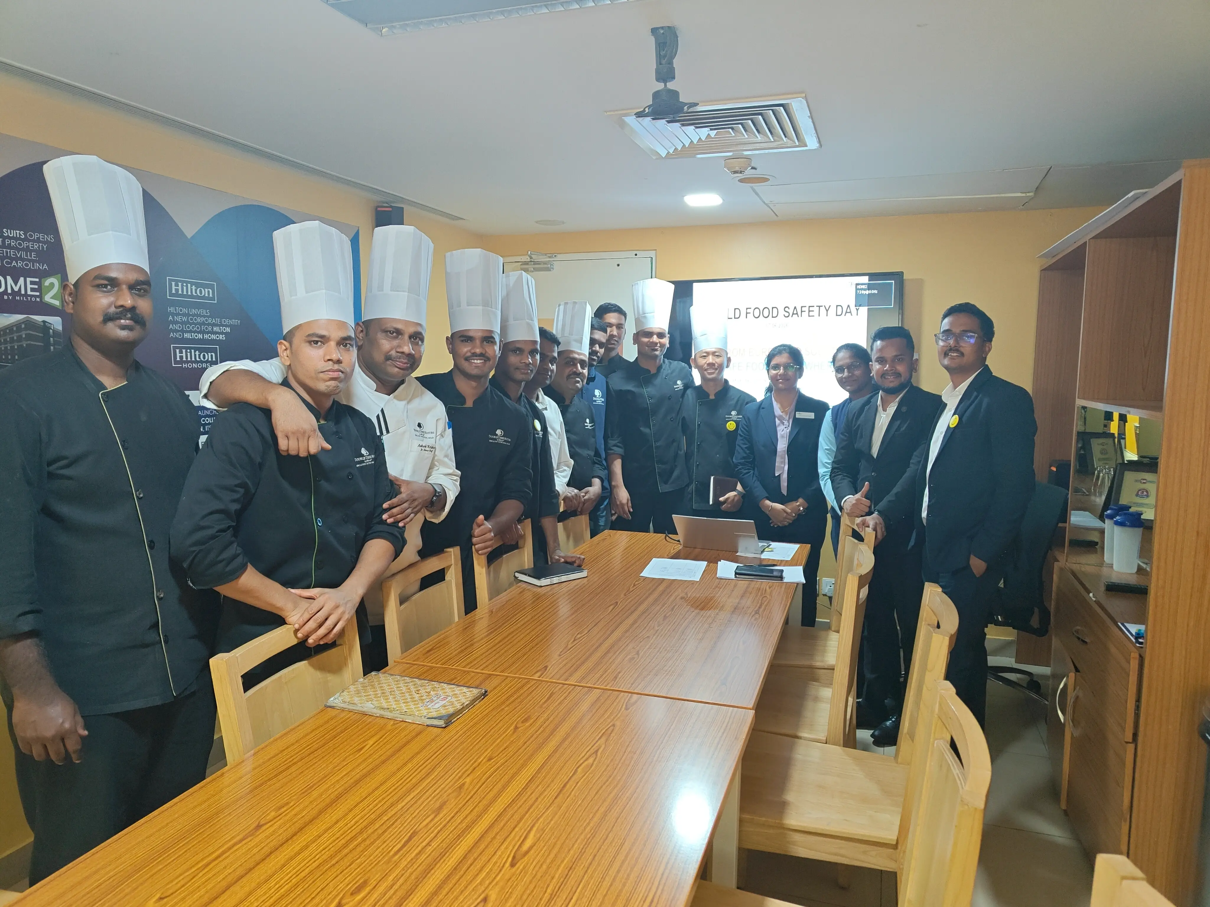 Chefs in white uniforms and hats standing with professionals in formal wear inside a boardroom before a training session.