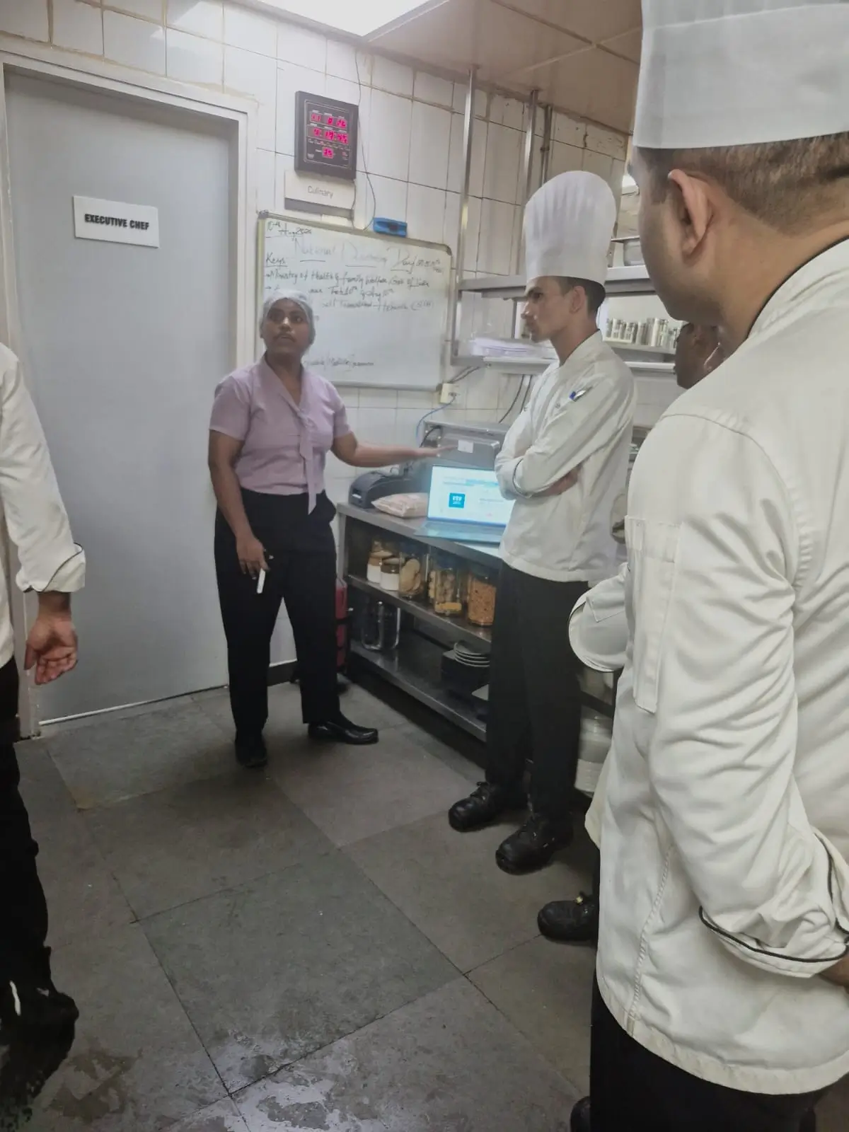 Chefs and an inspector checking stored ingredients on shelving inside a kitchen store room.